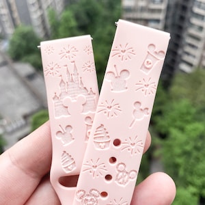 May include: Pink silicone watch band with a Disney theme. The band features a repeating pattern of Mickey Mouse heads, fireworks, and other Disney icons. The band is designed for a smartwatch.