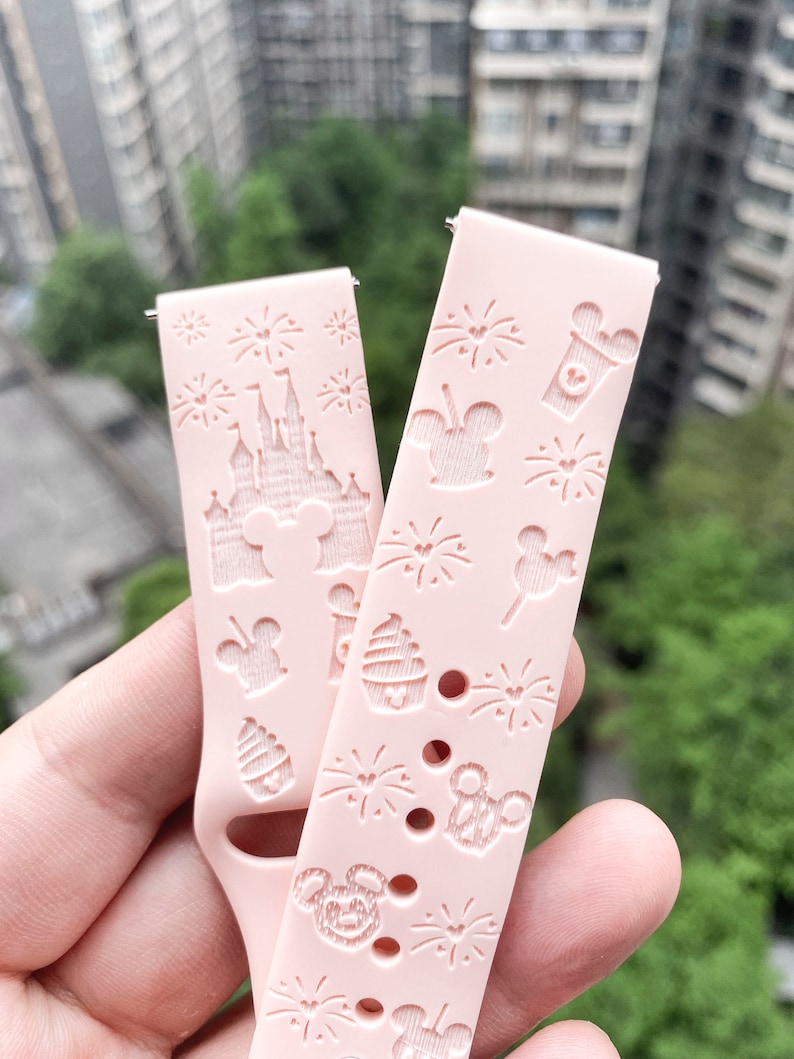 May include: Pink silicone watch band with a Disney theme. The band features a repeating pattern of Mickey Mouse heads, fireworks, and other Disney icons. The band is designed for a smartwatch.