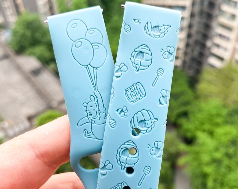 May include: Two light blue watch bands with different designs. One band features Winnie the Pooh holding balloons and the other band has a honeycomb and bee pattern.