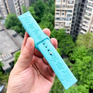 Puede incluir: Una correa de reloj de silicona azul claro con un patr&oacute;n repetido de tortugas marinas en relieve en la superficie.