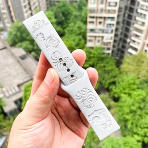 May include: A light gray watch band featuring engraved images of the Cheshire Cat from Alice in Wonderland. The band has a series of the cat's face and body. The band is held in a hand against a background of green trees and buildings.