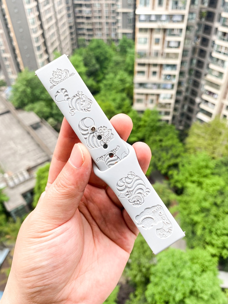 May include: A light gray watch band featuring engraved images of the Cheshire Cat from Alice in Wonderland. The band has a series of the cat's face and body. The band is held in a hand against a background of green trees and buildings.