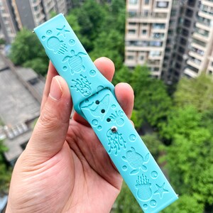 以下が含まれることがあります： 海の生き物のデザインが施された薄い青色のシリコン製腕時計バンド。バンドには、ウミガメ、ヒトデ、クラゲ、海藻が描かれています。