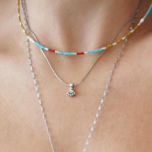 May include: A layered necklace set. The set includes a beaded choker in turquoise, red, and yellow, a silver pendant necklace, and a long silver lariat necklace with a small pendant and a dangling bead. Worn over a dark blue vest top.
