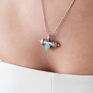 May include: A silver necklace with a cylindrical pendant and a small turquoise stone. The pendant is suspended from a delicate chain. The person is wearing a white strapless top.