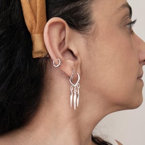 May include: Silver earrings with a dangling design. The earrings feature a small hoop and a larger earring with a heart-shaped top and three elongated, pointed pendants. A yellow headband is visible in the background.