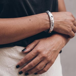 May include: A silver-toned metal bangle bracelet featuring a hammered, textured surface. The bracelet is adorned with small, circular decorative accents spaced evenly along the band, each containing a central dot surrounded by a beaded border.