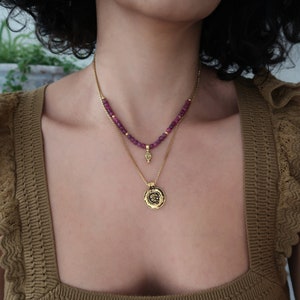 Op de afbeelding: Een gelaagde gouden ketting met een klein hangertje en een grotere ronde hanger. De ketting is gemaakt van kleine, donkerpaarse kralen.