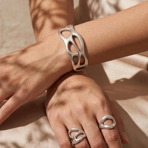 Peut inclure: Un bracelet manchette argenté avec un motif ajouré organique, accompagné de deux bagues argentées assorties présentant des formes fluides et irrégulières similaires. Les bijoux ont une finition lisse et polie au design continu et fluide.