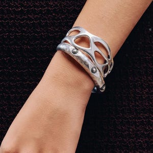 Peut inclure: Ensemble de bijoux argentés comprenant un bracelet manchette ajouré et une bague assortie. Un deuxième bracelet, plus simple, est également visible. Les bijoux sont portés sur le bras d'une personne, sur fond marron foncé.
