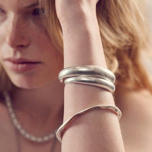 Könnte beinhalten: Eine Person trägt ein weißes Tanktop und Silberschmuck, darunter eine Halskette und mehrere Armbänder. Die Armbänder sind dick und scheinen aus Metall zu sein. Die Person berührt ihr Haar mit der Hand.