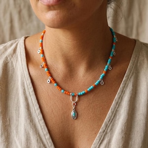 May include: A beaded necklace featuring alternating bright orange and turquoise blue beads. The necklace includes small silver-toned circular charms spaced along the strand and a central silver-toned pendant with a small turquoise inlay.