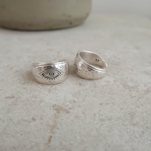 Peut inclure: Deux bagues argent&eacute;es &agrave; la texture martel&eacute;e. L'une des bagues pr&eacute;sente un symbole d'&oelig;il grav&eacute; en noir avec des lignes rayonnantes. L'autre bague est ouverte et affiche le nombre 17 grav&eacute; sur la surface int&eacute;rieure de l'anneau.