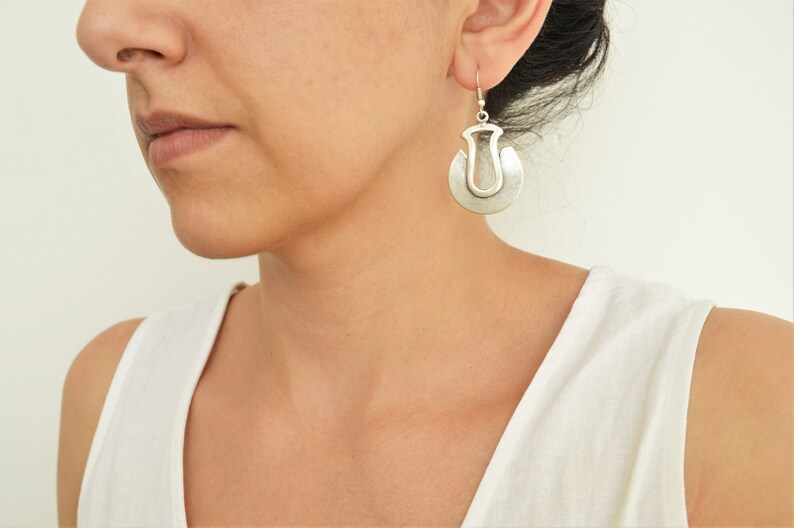 May include: Silver dangle earrings with a unique, abstract design. The earrings are made of two overlapping circles, one with a cutout in the shape of a teardrop.