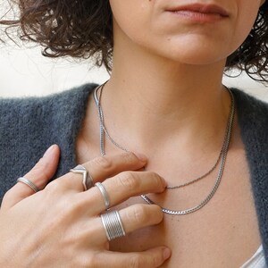以下が含まれることがあります： 女性が2本のチェーンのシルバーネックレスをしています。彼女はまた、指にいくつかのシルバーリングをしています。