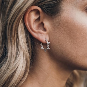 Op de afbeelding: Een kleine zilveren oorring met een reeks gelijkmatig verdeelde, kleine bolvormige kralen langs de buitenrand. De oorring heeft een gepolijste metalen afwerking en een eenvoudig scharnierend sluitingsmechanisme in een cirkelvorm.