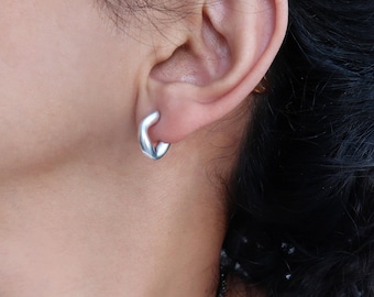 May include: A close-up of a person's ear with a silver hoop earring. The earring is a simple, modern design with a slightly curved shape.