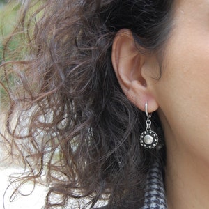 May include: A silver hoop earring with a small, round, black and silver charm. The charm has a textured, circular design.