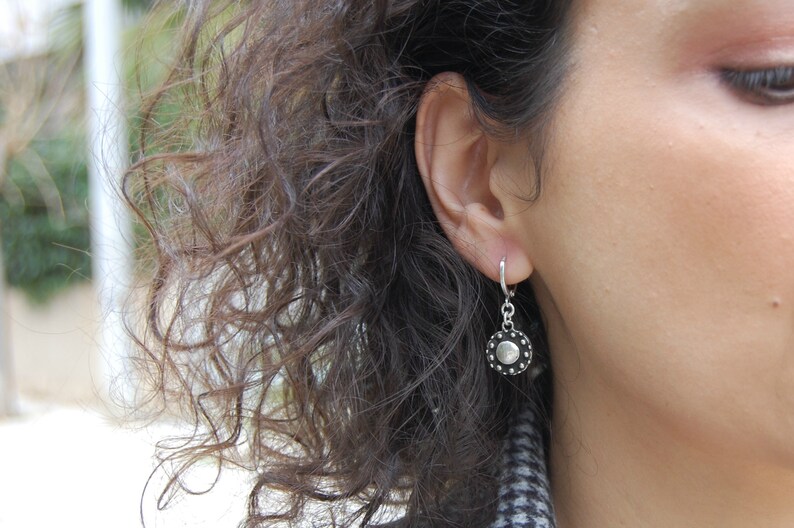 May include: A silver hoop earring with a small, round, black and silver charm. The charm has a textured, circular design.