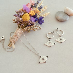 May include: A silver necklace and earrings set with a circular pendant and matching earrings. The pendant and earrings have a textured, hammered look and feature a small circle in the centre. The jewellery is displayed on a white surface with a dried flower bouquet and two grey stones.