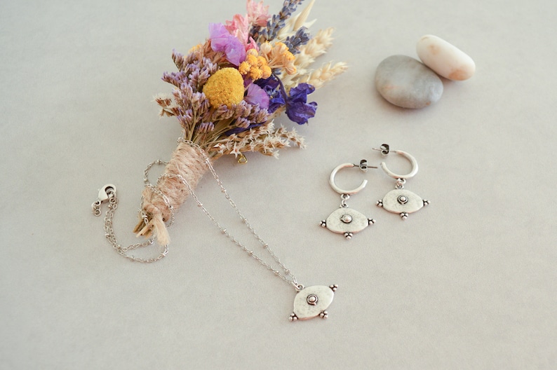 May include: A silver necklace and earrings set with a circular pendant and matching earrings. The pendant and earrings have a textured, hammered look and feature a small circle in the centre. The jewellery is displayed on a white surface with a dried flower bouquet and two grey stones.