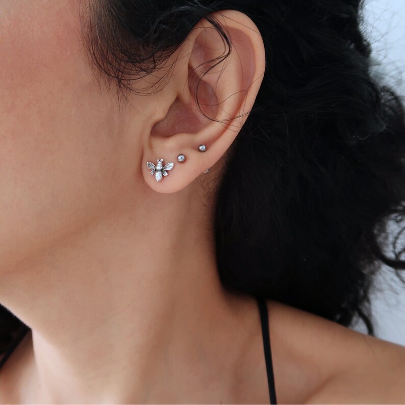 May include: A close-up of a person's ear with three earrings. The bottom earring is a silver bee with clear crystals. The middle earring is a small silver ball. The top earring is a small silver ball.