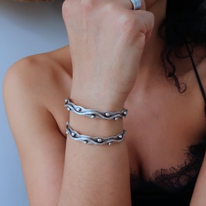 Puede incluir: Dos pulseras de plata con un diseño ondulado y pequeñas cuentas redondas. Las pulseras están apiladas en la muñeca de una persona.