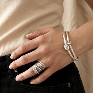 May include: A silver-toned ring and a matching silver-toned bracelet featuring a textured, organic surface. The ring has a wide band with diagonal ridges, while the bracelet consists of two thin, interconnected bands with a knotted design.