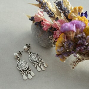 Puede incluir: Pendientes de plata con un dise&ntilde;o circular de filigrana y una perla en la parte superior. Los pendientes se muestran sobre una superficie blanca junto a un ramo de flores secas.