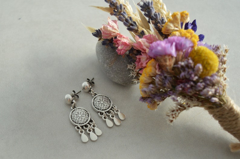 Puede incluir: Pendientes de plata con un dise&ntilde;o circular de filigrana y una perla en la parte superior. Los pendientes se muestran sobre una superficie blanca junto a un ramo de flores secas.