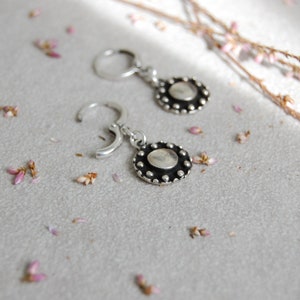 May include: A pair of silver hoop earrings with a round, textured silver charm hanging from each hoop.
