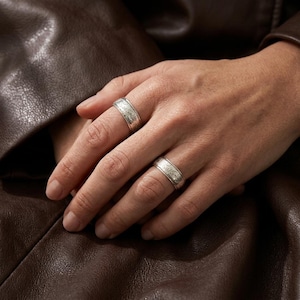 May include: Two silver-toned metal rings featuring an intricate, engraved floral pattern across the band. Each ring has a polished finish with subtle detailing along the edges, worn on the index and ring fingers of a hand against a dark brown leather.