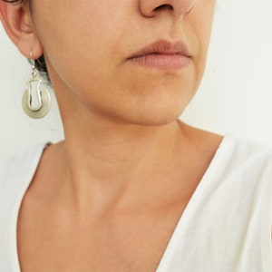 May include: A woman wearing a white tank top with silver hoop earrings. The earrings have a unique design with a cutout shape.