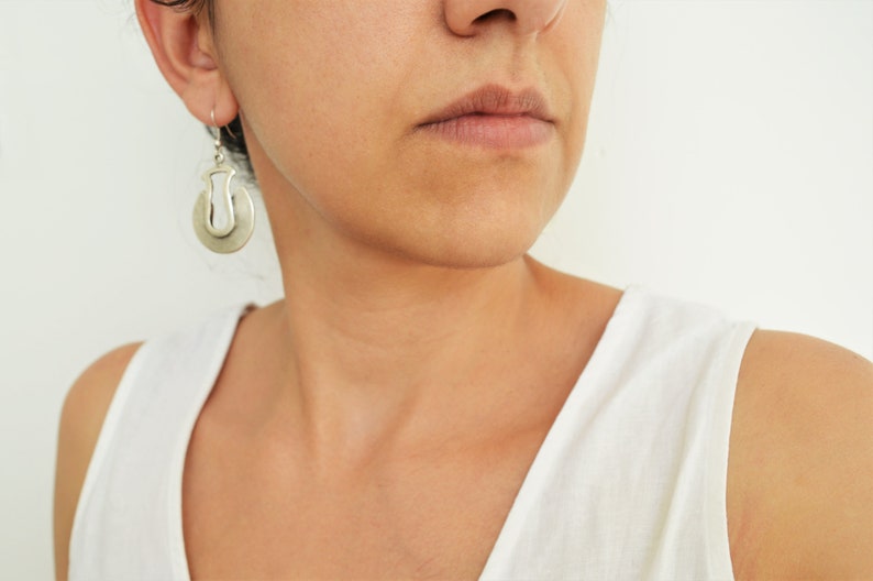 May include: A woman wearing a white tank top with silver hoop earrings. The earrings have a unique design with a cutout shape.