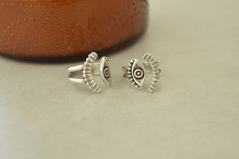 May include: Two silver toe rings with an eye design. The rings are open and adjustable.
