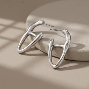 Op de afbeelding: Een paar zilverkleurige oorbellen met een onregelmatig, open frameontwerp en een gladde, gepolijste afwerking. Elke oorbel heeft een stekersluiting en een vloeiende vorm die een holle, driehoekige silhouet creëert.