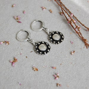 May include: A pair of silver hoop earrings with a small, round, silver charm hanging from each hoop. The charms have a textured, circular design with a raised center.