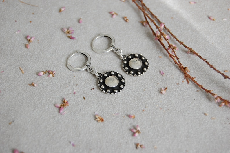 May include: A pair of silver hoop earrings with a small, round, silver charm hanging from each hoop. The charms have a textured, circular design with a raised center.