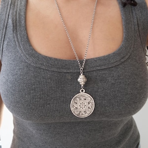 May include: Silver-toned pendant necklace with a long chain. The pendant features an ornate, circular design with a central square and swirling patterns. The necklace is worn over a gray tank top.