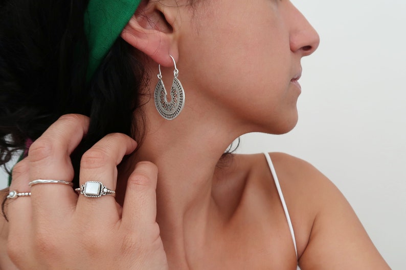 Puede incluir: Pendientes plateados, de dise&ntilde;o intrincado con forma de l&aacute;grima. Los pendientes presentan un patr&oacute;n calado detallado. Tambi&eacute;n se ve un anillo plateado con una piedra blanca cuadrada. La imagen muestra joyas.