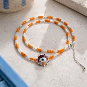 May include: A beaded necklace featuring a central circular pendant with an eye motif in shades of maroon, white, light blue, and orange. The strand consists of small orange, white, and light blue beads with a silver-toned metal lobster clasp and chain.