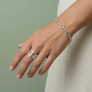 Può includere: Un anello color argento caratterizzato da una fascia larga con un intaglio centrale a forma di lettera D maiuscola, abbinato a un bracciale a catena color argento composto da maglie circolari e piccoli dettagli a forma di D con chiusura.