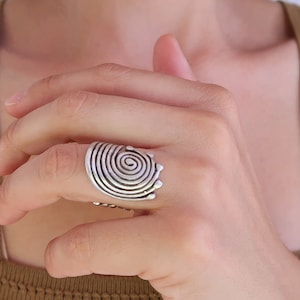 Op de afbeelding: Een zilveren ring met een spiraalvormig ontwerp. De ring wordt aan een vinger gedragen.