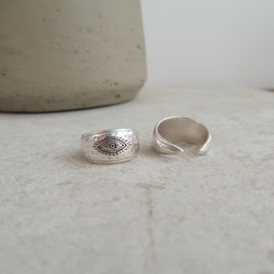Peut inclure: Deux bagues argent&eacute;es &agrave; la texture martel&eacute;e. L'une des bagues pr&eacute;sente un symbole d'&oelig;il grav&eacute; avec des lignes rayonnantes, tandis que l'autre est une bague ouverte aux extr&eacute;mit&eacute;s lisses et effil&eacute;es. Les deux pi&egrave;ces ont une finition m&eacute;tallique.