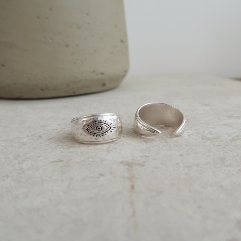Peut inclure: Deux bagues argent&eacute;es &agrave; la texture martel&eacute;e. L'une des bagues pr&eacute;sente un symbole d'&oelig;il grav&eacute; avec des lignes rayonnantes, tandis que l'autre est une bague ouverte aux extr&eacute;mit&eacute;s lisses et effil&eacute;es. Les deux pi&egrave;ces ont une finition m&eacute;tallique.