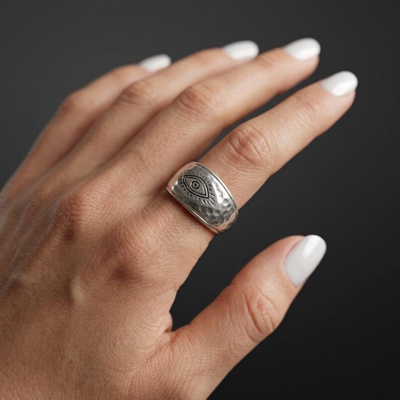 Peut inclure: Une large bague en m&eacute;tal argent&eacute; avec une texture martel&eacute;e et un motif d'&oelig;il grav&eacute; au centre. Le design comprend des lignes rayonnantes autour de l'&oelig;il, cr&eacute;ant un point focal d&eacute;taill&eacute; sur la surface polie du m&eacute;tal de la bague.