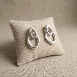 May include: A pair of silver-toned earrings featuring an abstract, organic shape. Each earring consists of two interconnected, irregular loops with a smooth, matte finish. The design is asymmetrical and sculptural, showcasing a fluid, open-frame structure.