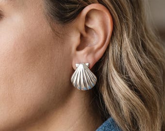 May include: A silver-toned earring shaped like a scallop shell with vertical ridges and a smooth, polished finish. The earring is secured to the earlobe with a post-style backing, featuring a rounded top edge and a fan-like base.