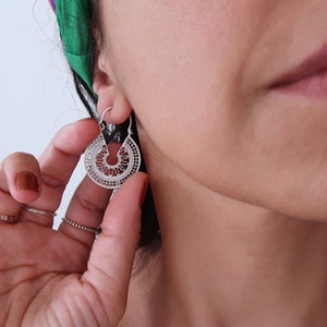 Puede incluir: Pendientes de aro de filigrana plateada con un dise&ntilde;o detallado y calado. Los pendientes tienen forma de semic&iacute;rculo con intrincados patrones, creando un aspecto delicado y elegante. Los pendientes se est&aacute;n usando.