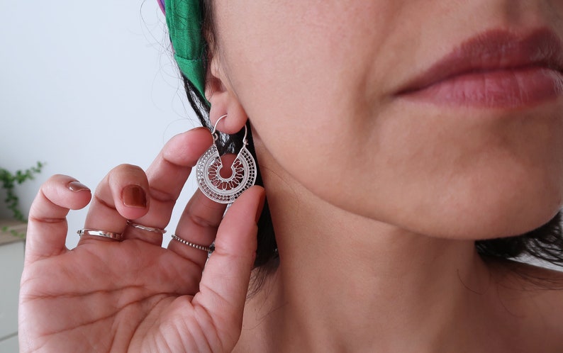 Puede incluir: Pendientes de aro de filigrana plateada con un dise&ntilde;o detallado y calado. Los pendientes tienen forma de semic&iacute;rculo con intrincados patrones, creando un aspecto delicado y elegante. Los pendientes se est&aacute;n usando.
